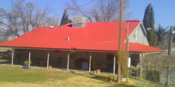 A house with a large, bright red metal roof, a wide porch with support posts, and a grassy lawn in the foreground.