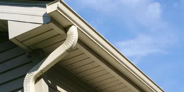 White rain gutter and downspout attached to the corner of a house with beige siding against a blue sky.