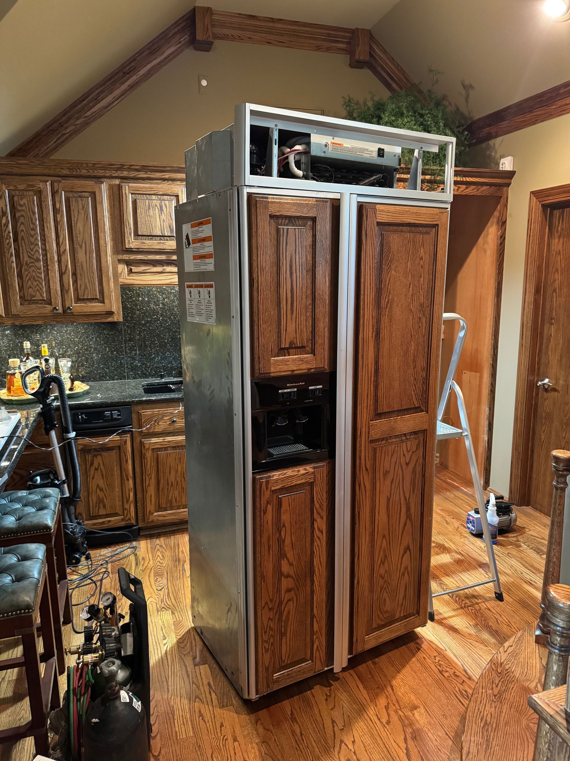 Large refrigerator, stainless steel with wood doors, in a kitchen setting. A ladder stands nearby.