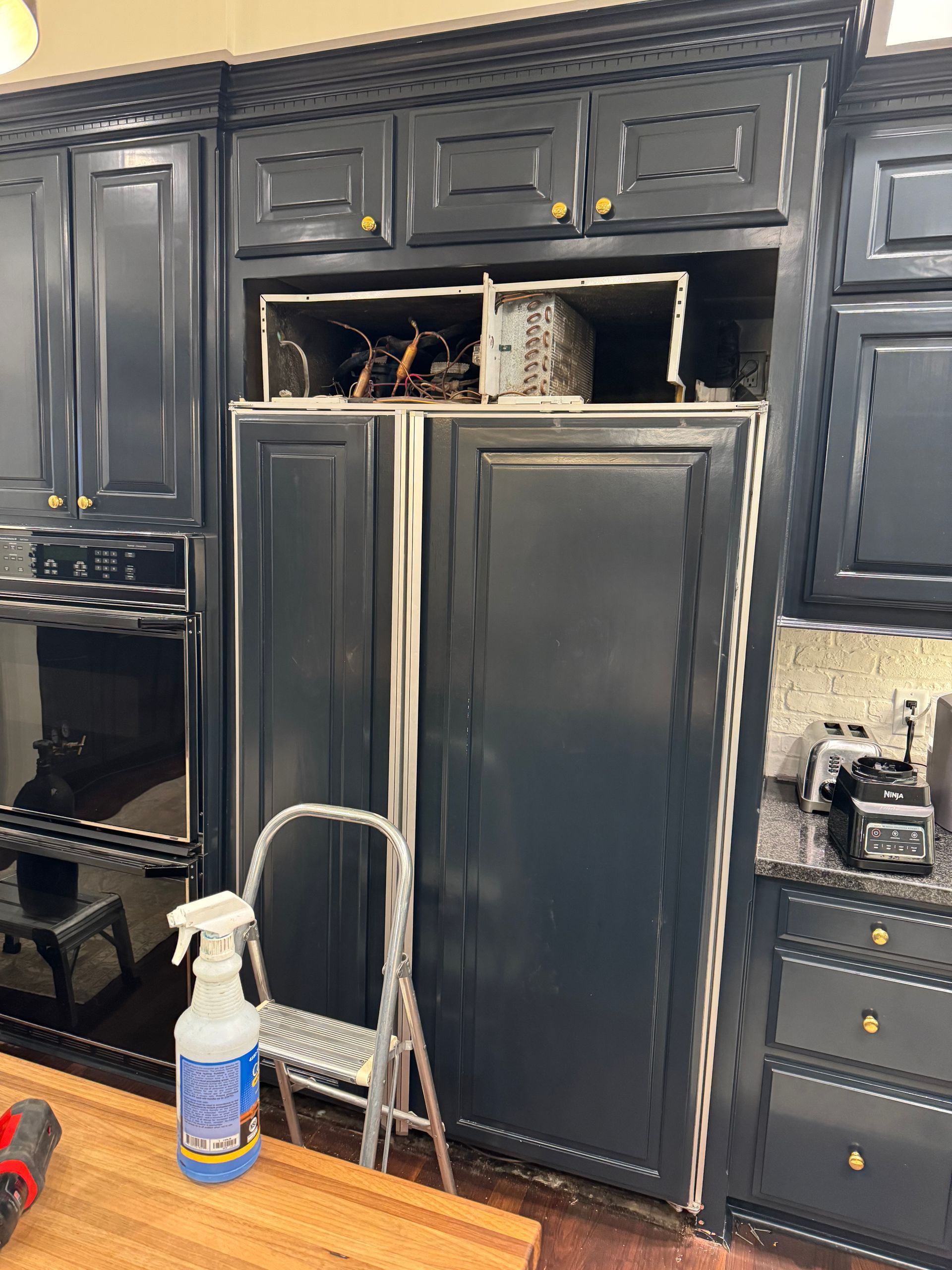 Refrigerator with upper cabinet removal, step stool, spray bottle, and surrounding dark blue kitchen cabinets.