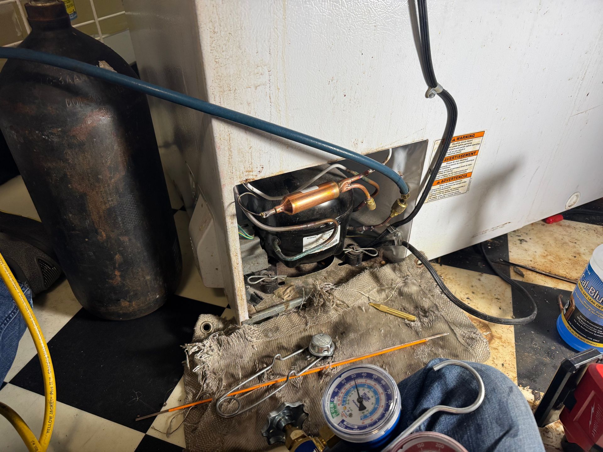 Refrigerator repair: Technician working on the compressor. Tools and gauges are visible with a gas cylinder nearby.