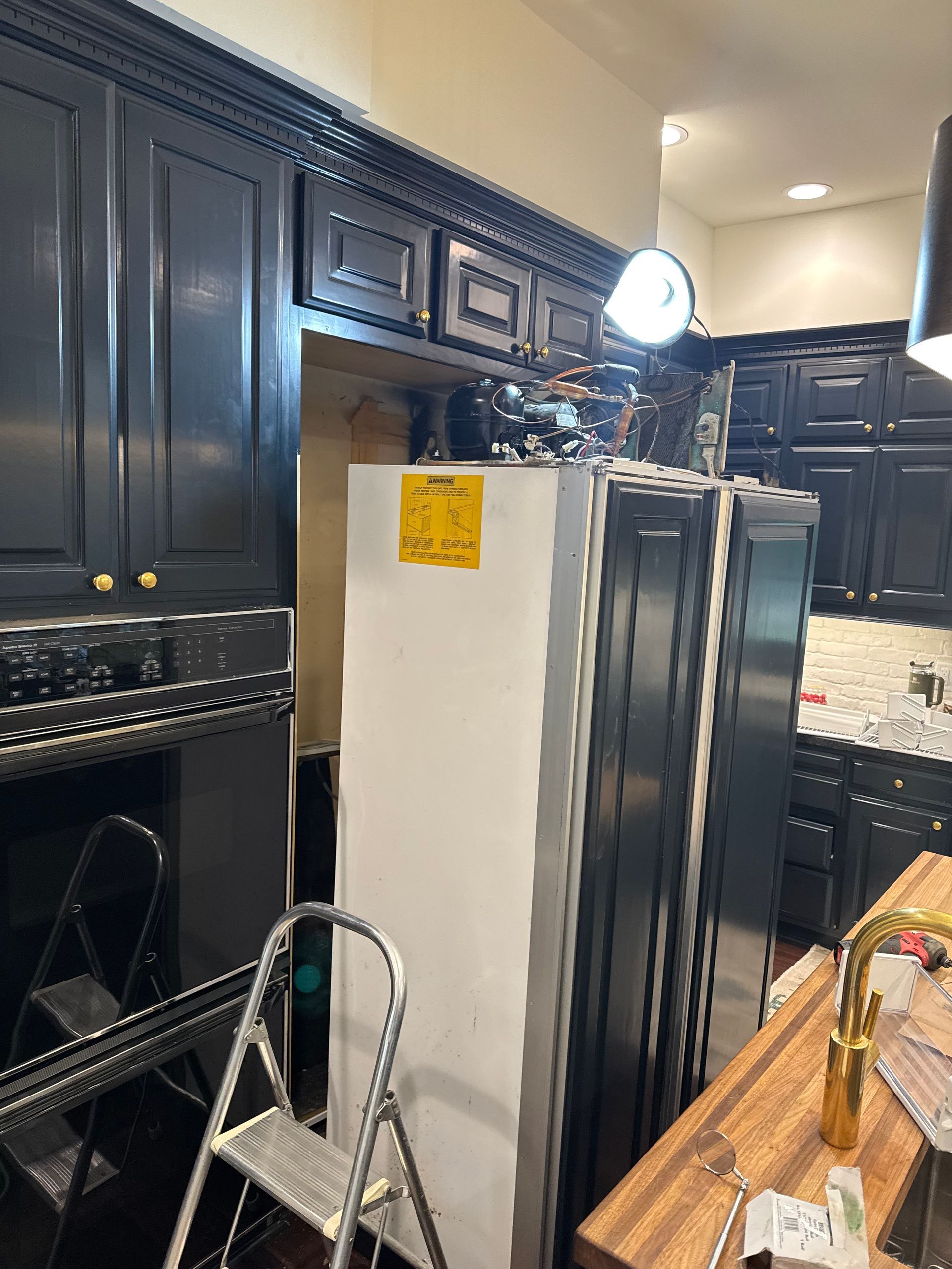 Refrigerator being moved into a kitchen with dark cabinets, a ladder, and a work light.