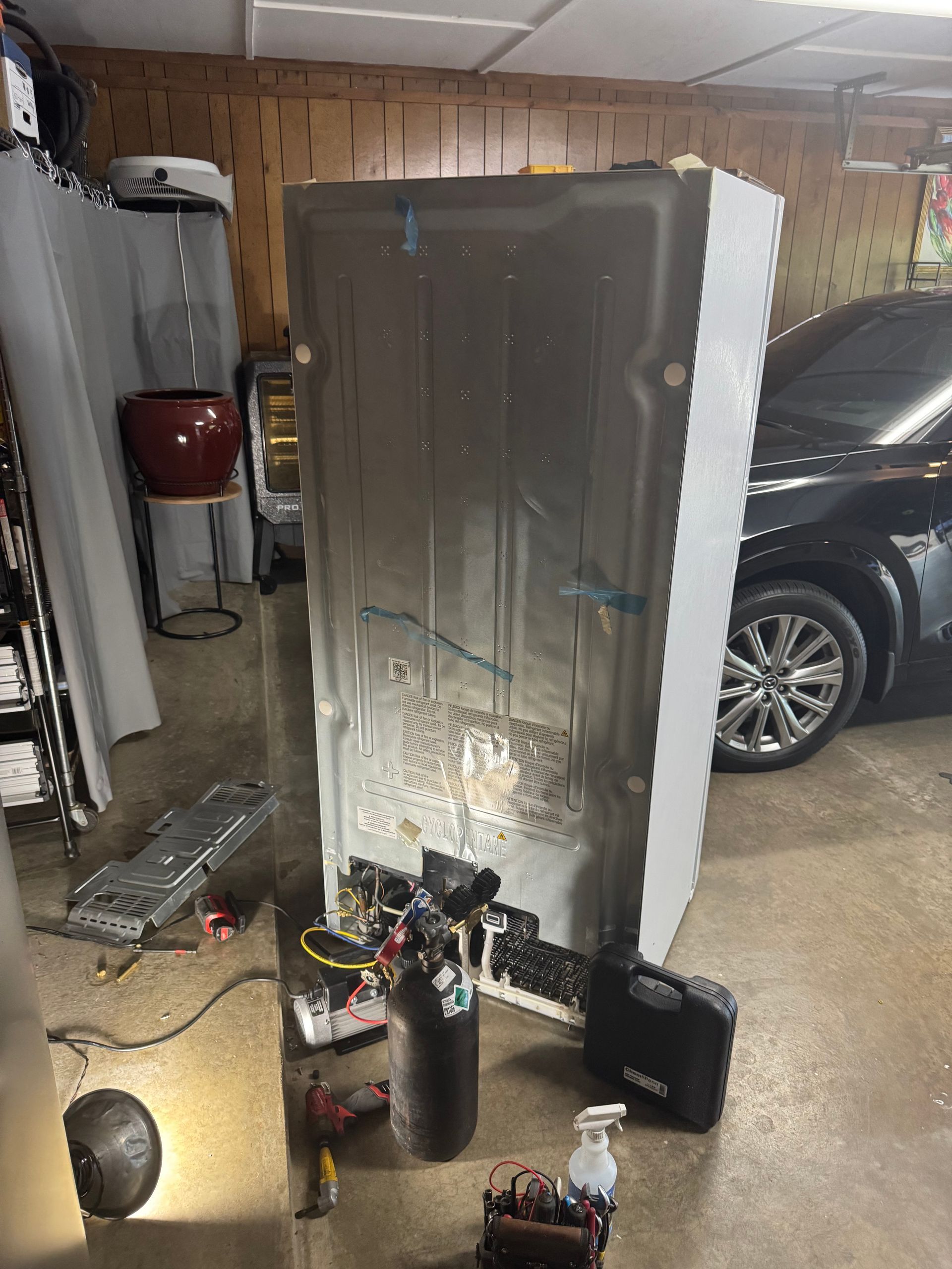 Refrigerator repair in a garage; rear view shows components, tools, and a black car.