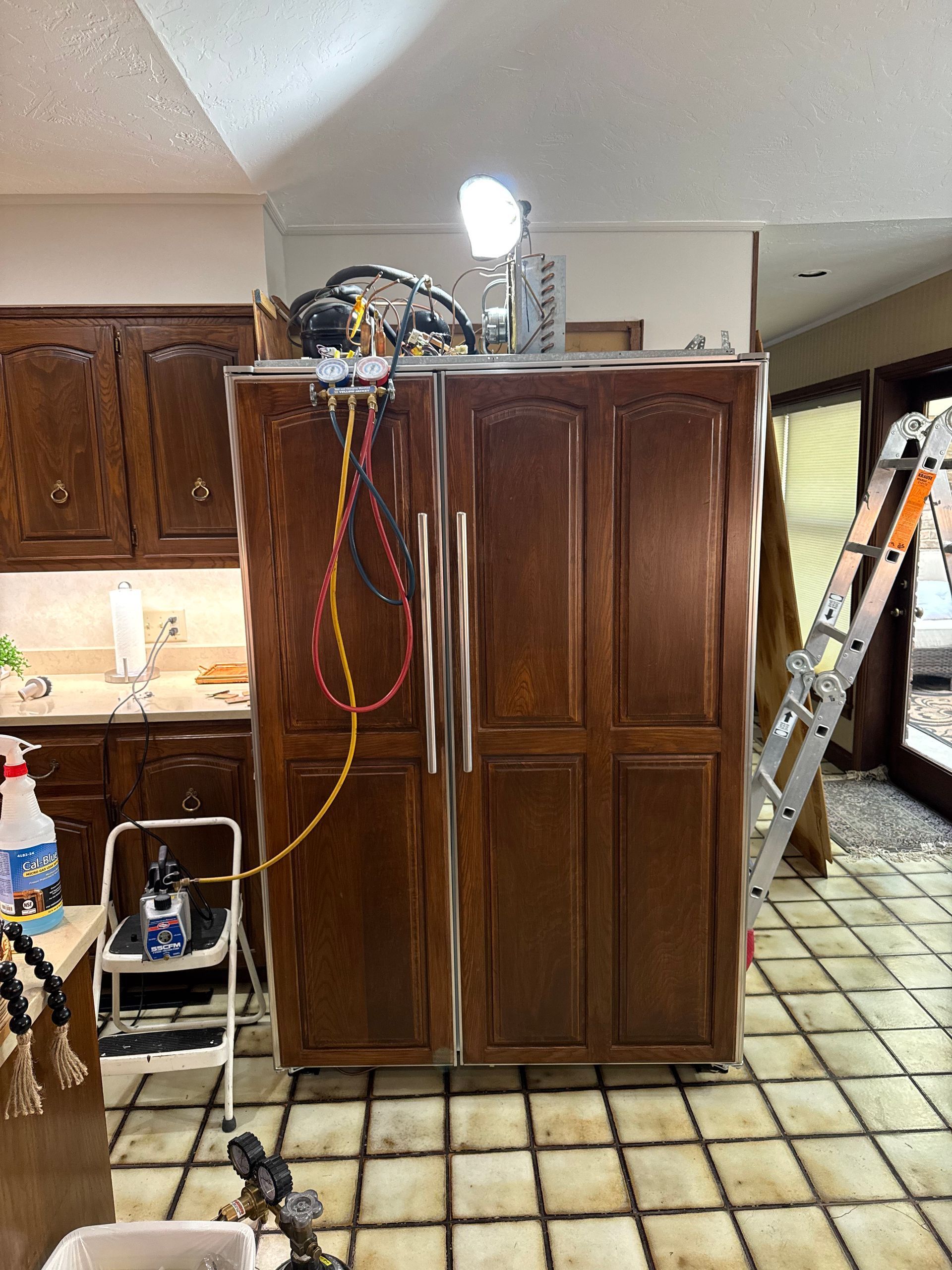 Refrigerator being serviced in a kitchen with brown cabinets and tiled floor. A ladder leans nearby.