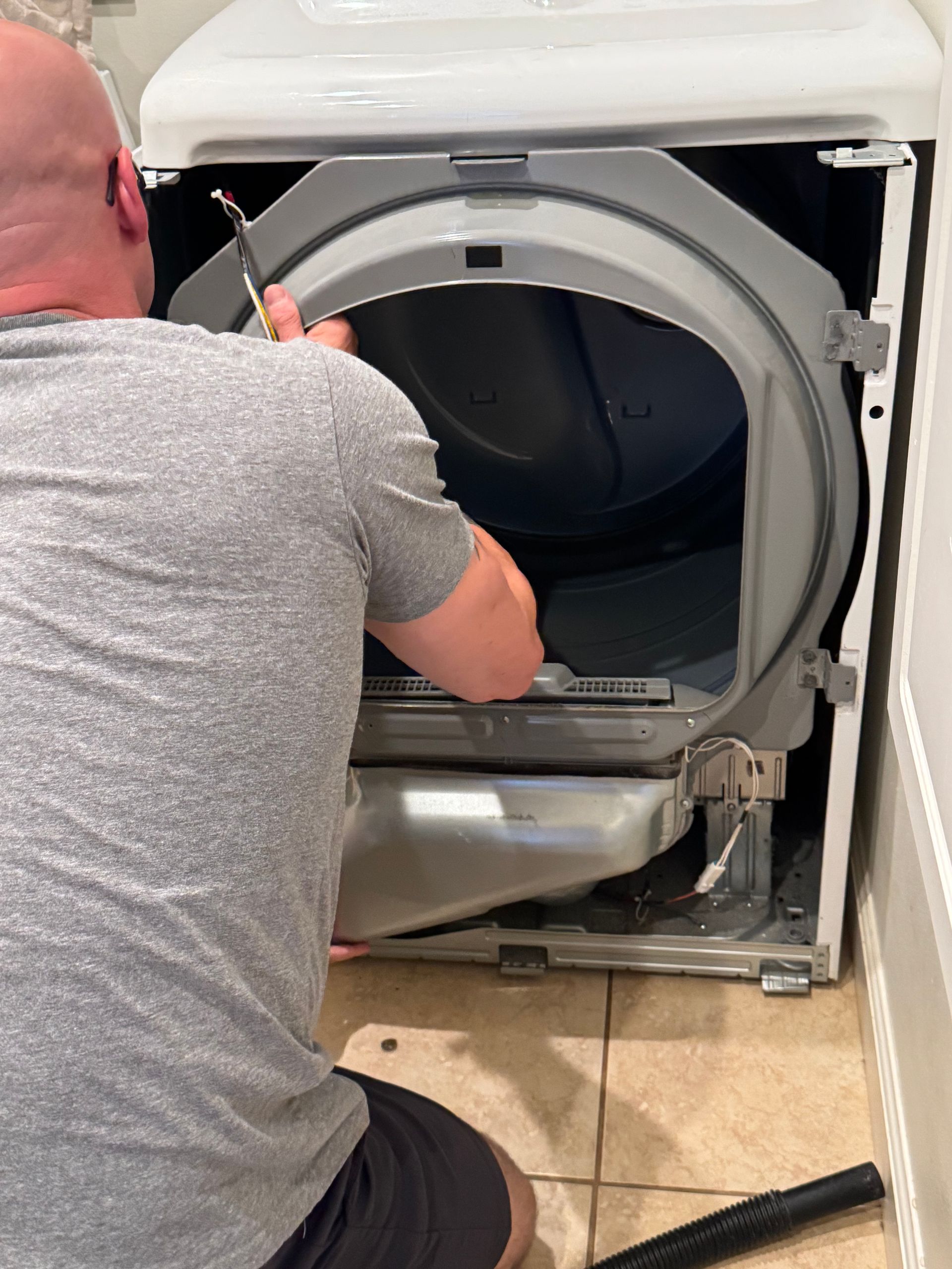 Person repairs dryer, removing debris with a tool. White appliance, tan tiled floor.