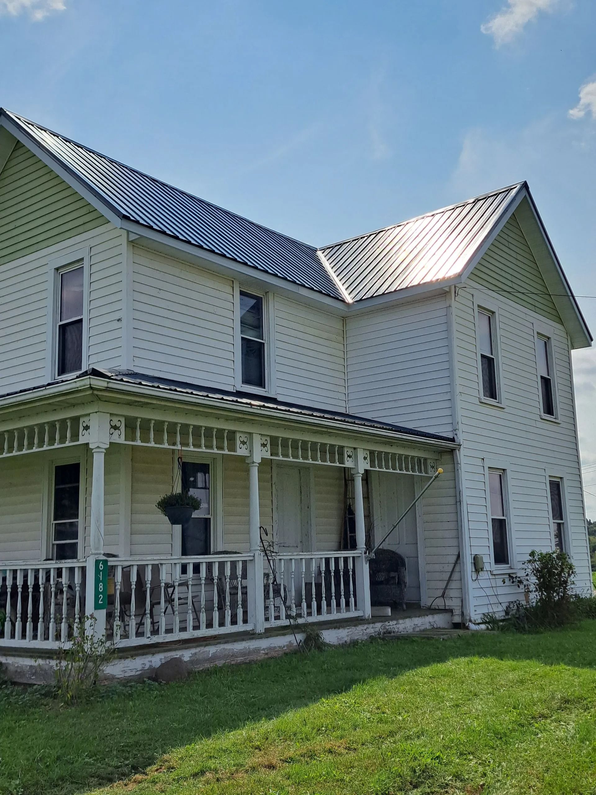 White two-story farmhouse with a dark roof and a porch. Green grass, blue sky.