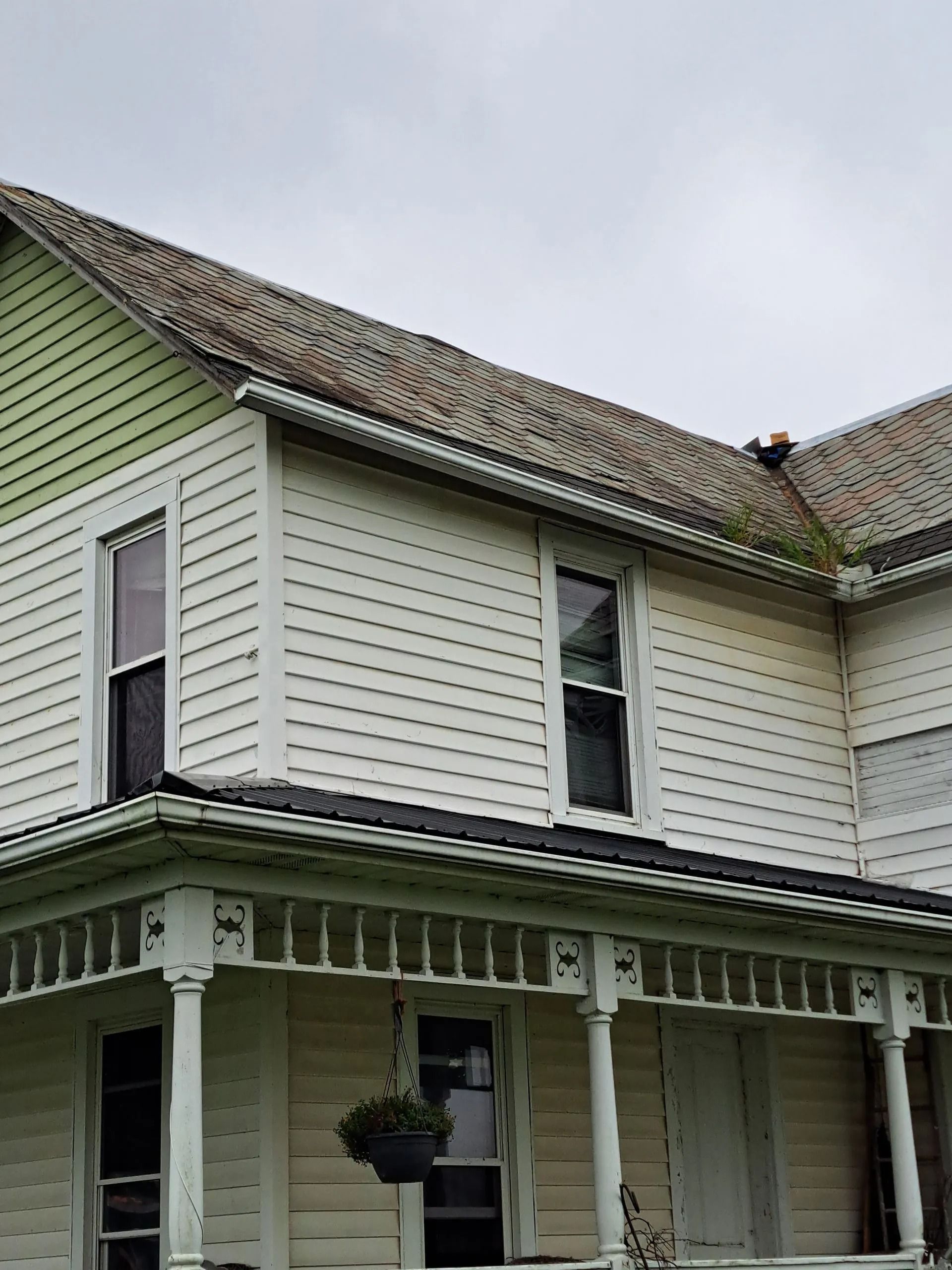 Two-story white house with a porch. Green roof with moss. Overcast sky.