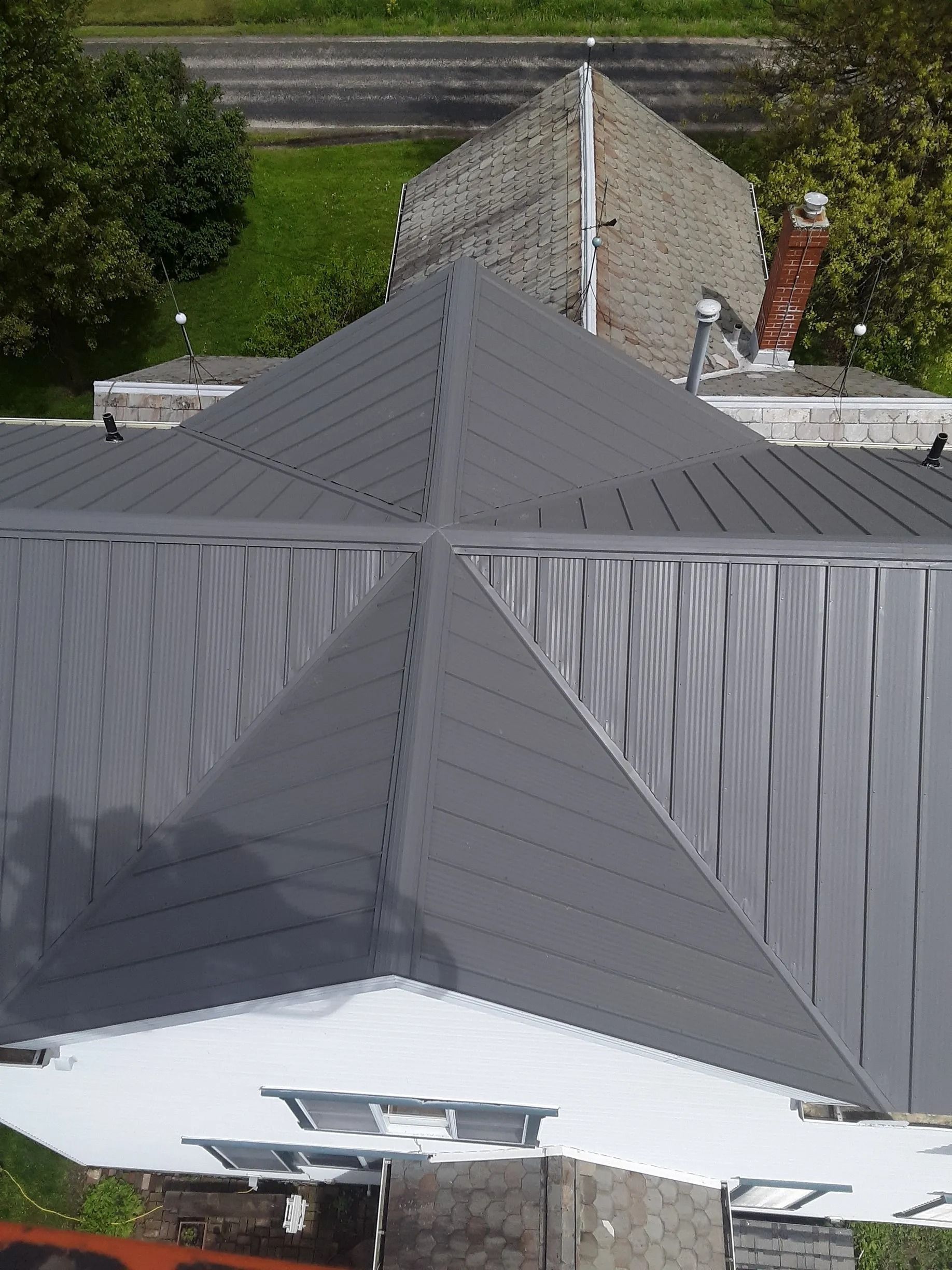 Gray metal roof of a building with triangular sections, set against a green lawn and a brick chimney.