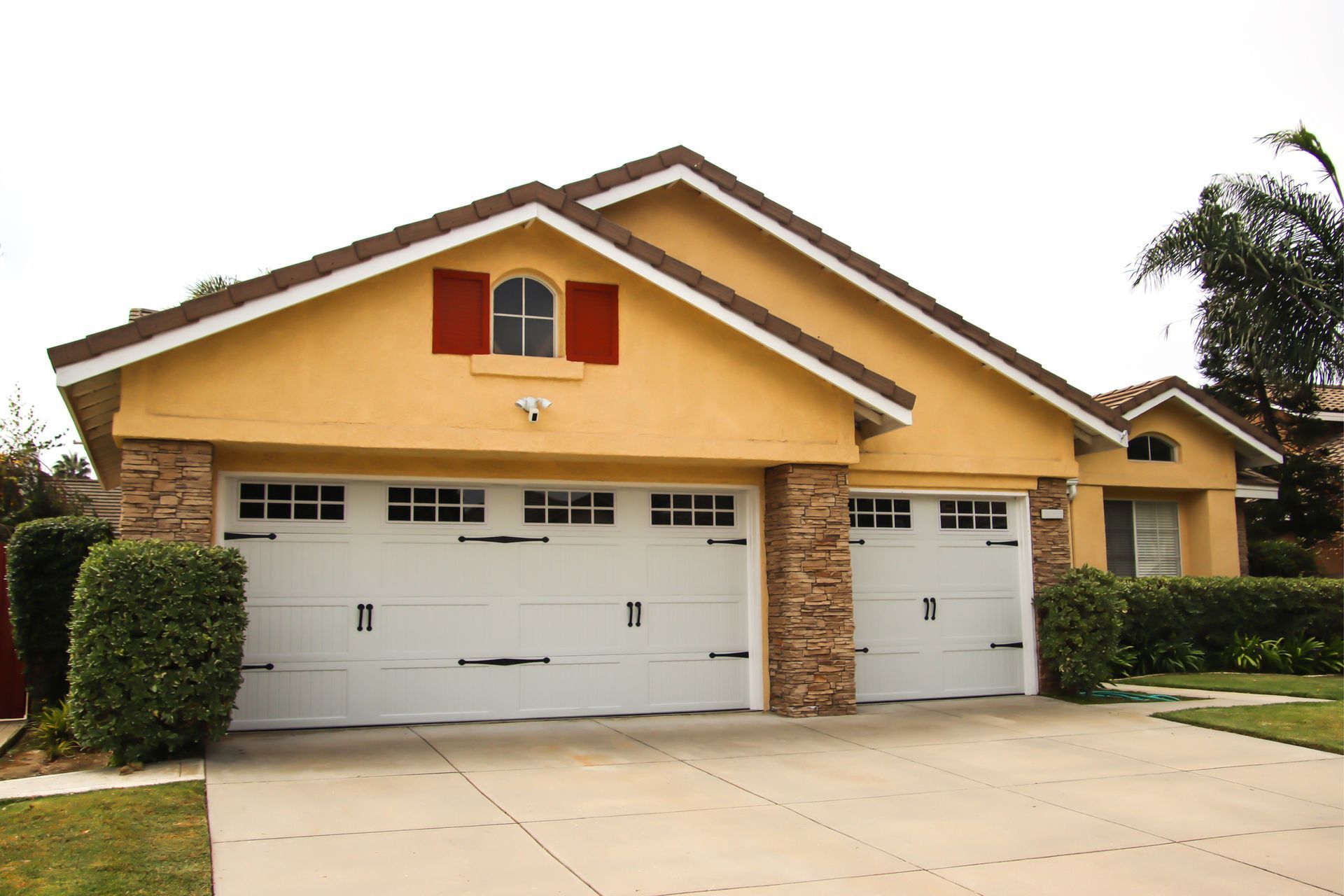 Yellow house with white garage doors, brown stone accents, and red shutters.