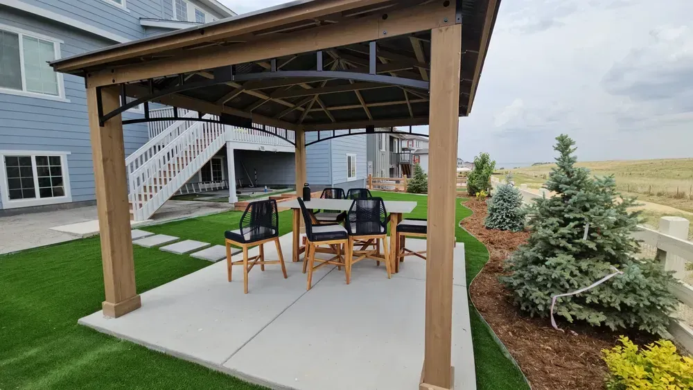 A gazebo with a dining set on a concrete patio, adjacent to a house and yard with trees.