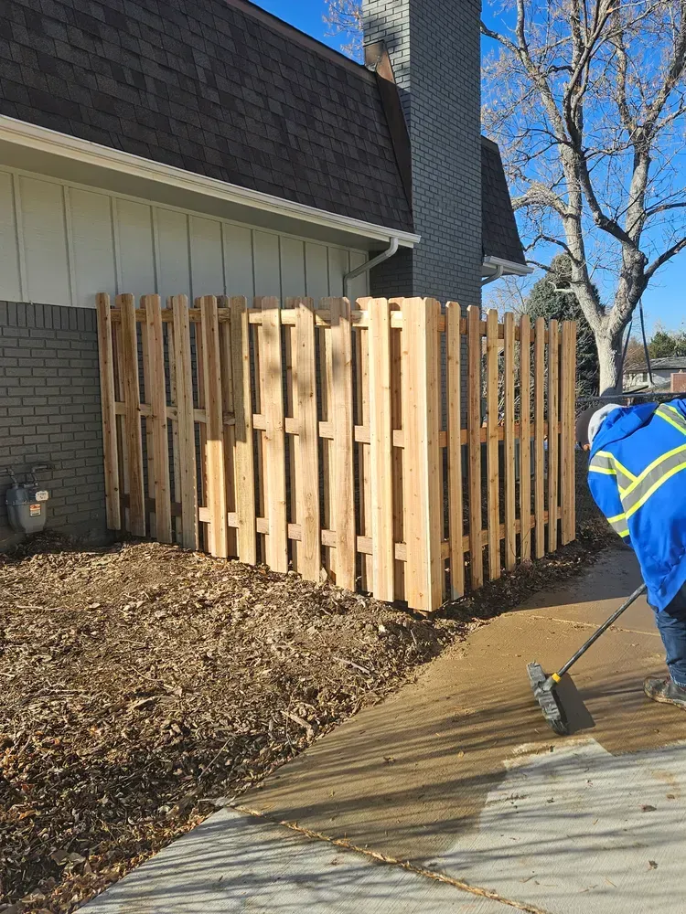 A wooden fence encloses utilities beside a house; a person sweeps a sidewalk.