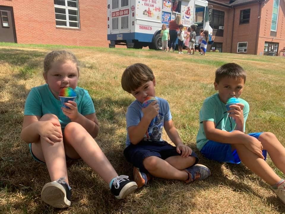 Three children are sitting on the grass eating ice cream in front of an ice cream truck.