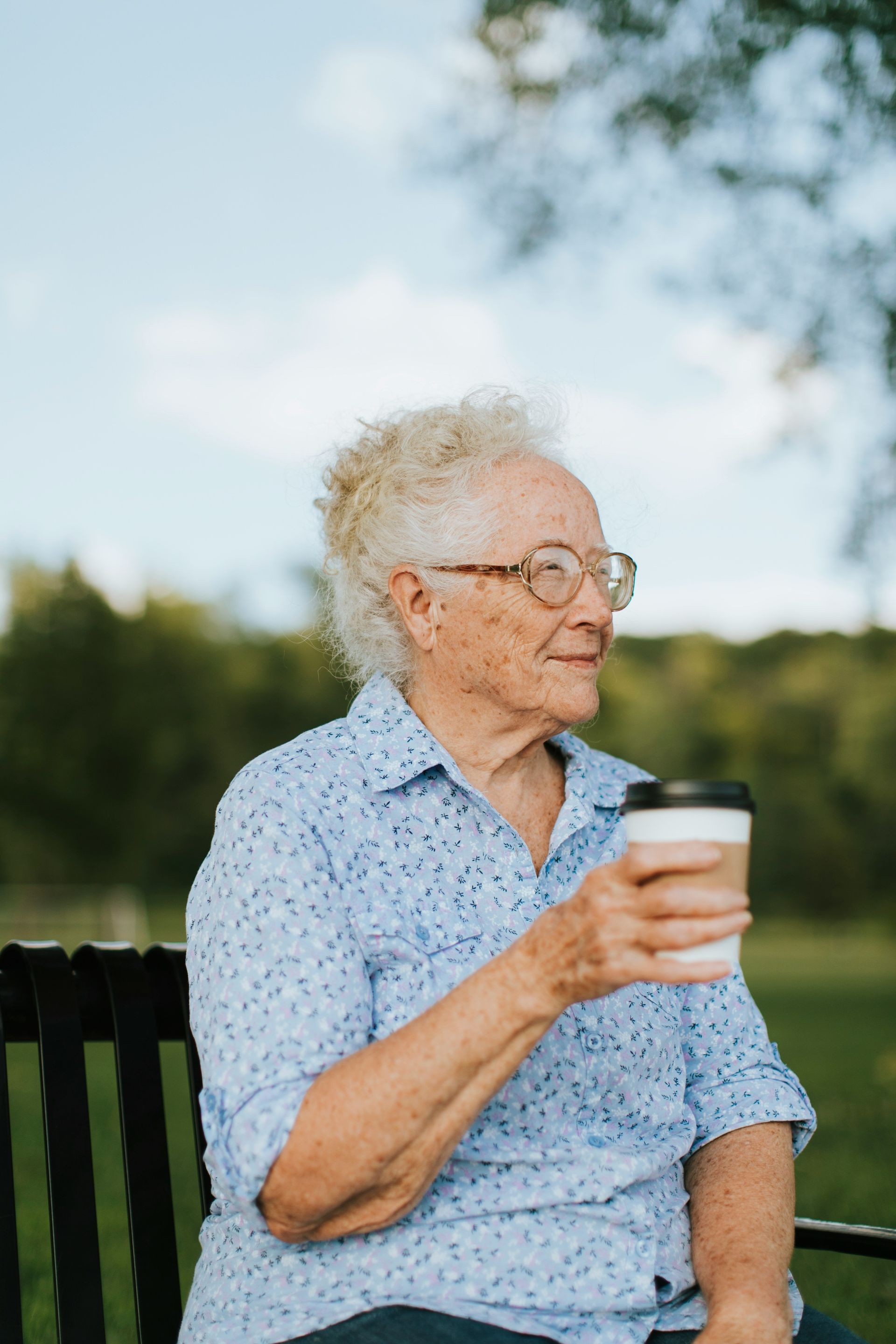 An elderly woman is sitting on a park bench holding a cup of coffee.