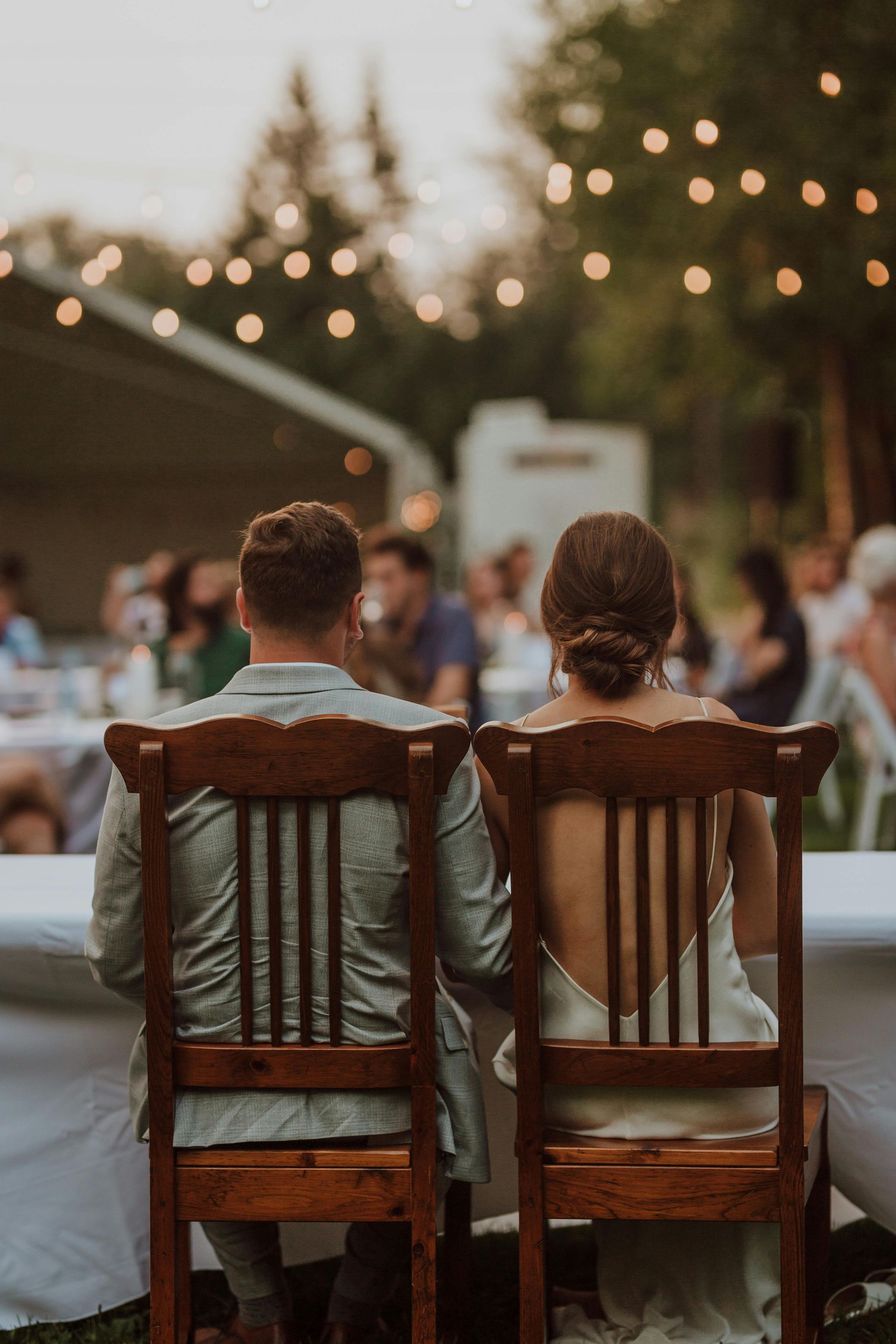 A bride and groom are sitting in wooden chairs at a wedding reception.