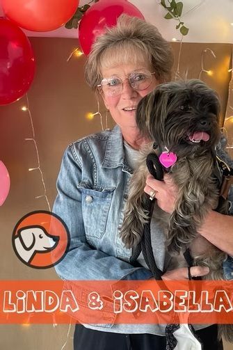 A person with glasses holding a small dog with a pink collar, in front of a backdrop with red balloons and fairy lights.