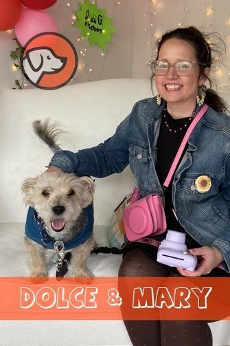 A smiling person with glasses and a denim jacket pets a small, scruffy dog in a blue harness against a festive background.