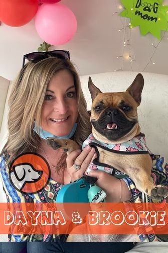 A smiling woman holds a fawn-colored French Bulldog wearing a patterned vest, with balloons and a 
