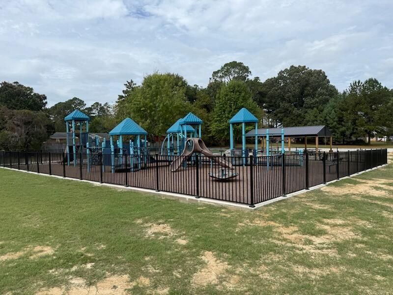 A fenced playground featuring blue play structures with slides and towers on a brown surface set against a grassy lawn.