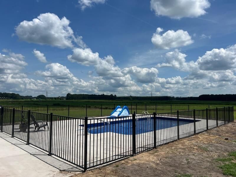 A blue rectangular swimming pool with a small slide, enclosed by a black metal fence against a blue, cloudy sky.