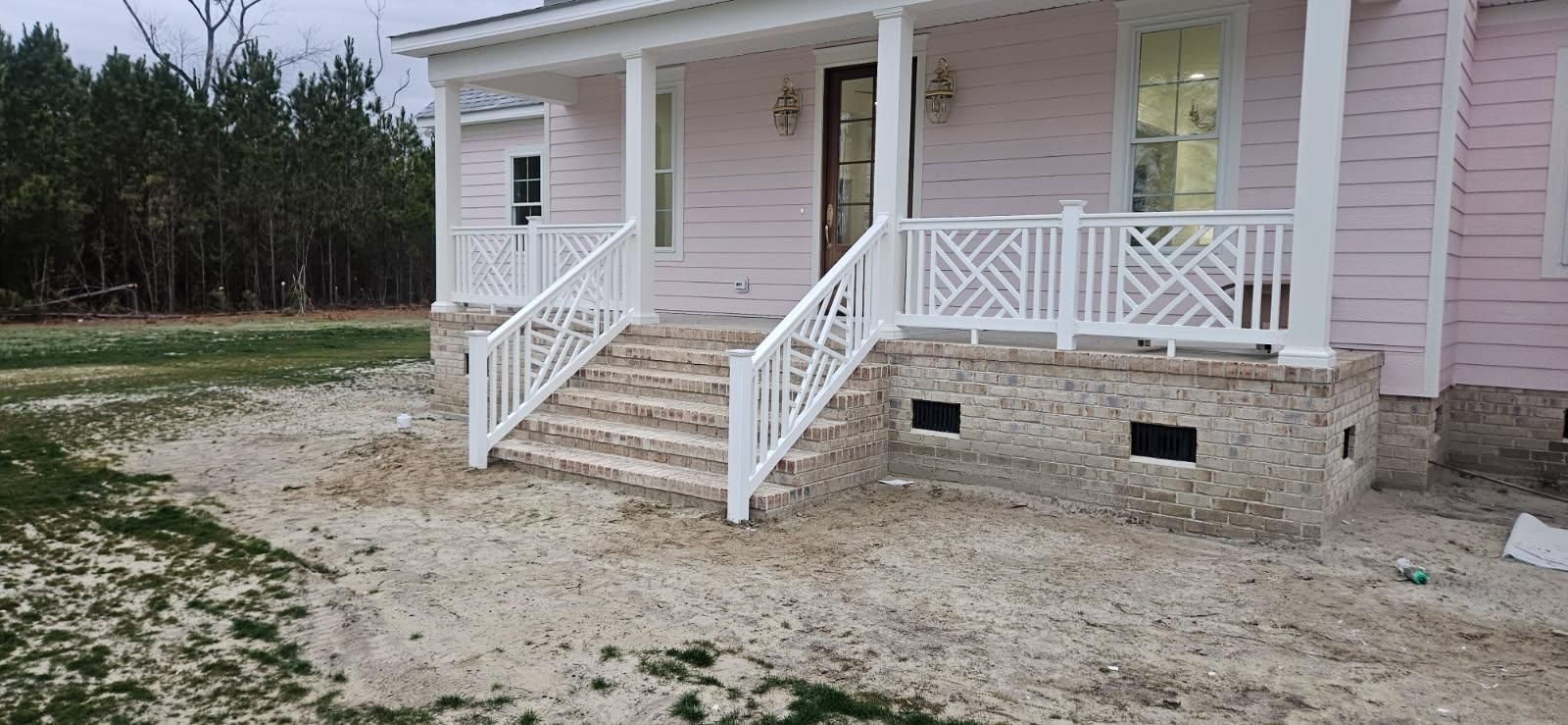 A pastel pink house with a stone-foundation porch, white railings, and wide front steps set against a grassy field.