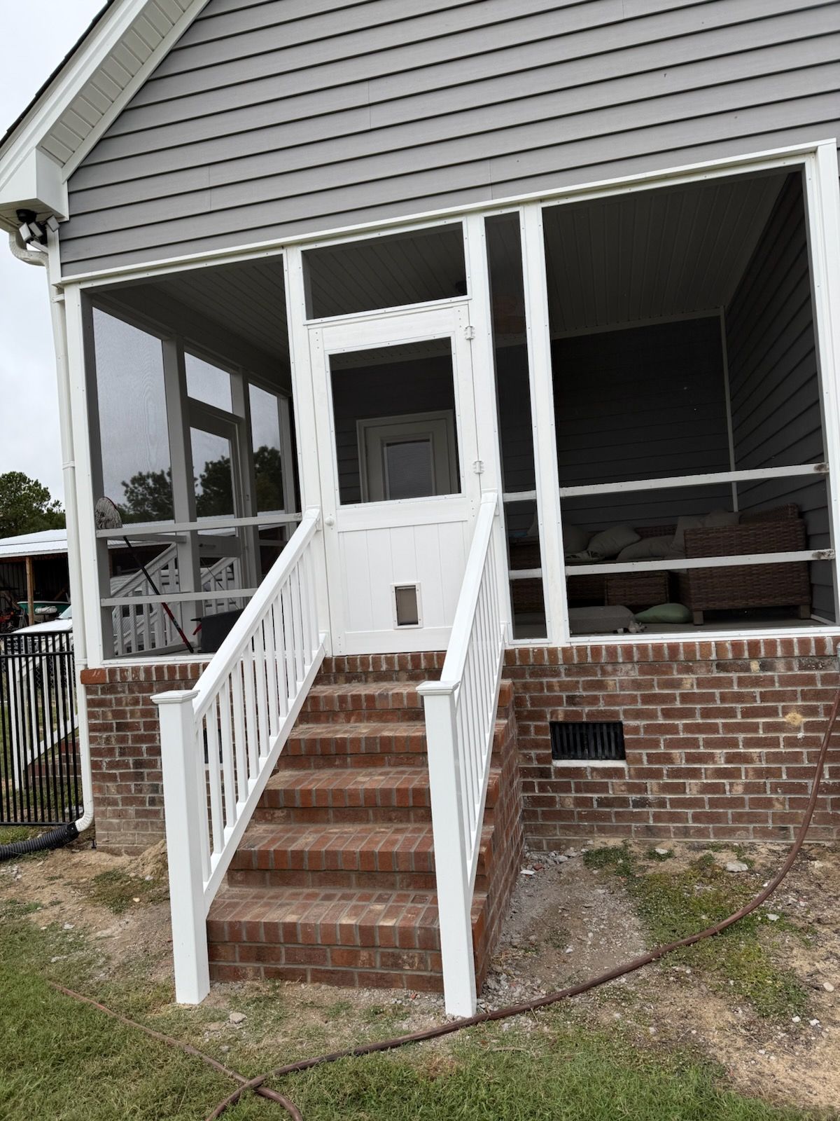 A screened-in porch with gray siding above a brick foundation, featuring a white door and stairs with white railings.