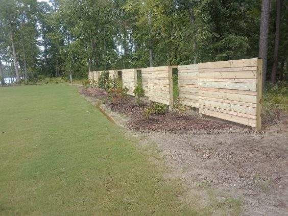 A horizontal wood-slat fence stands along a mulched garden bed next to a grassy lawn, with trees in the background.
