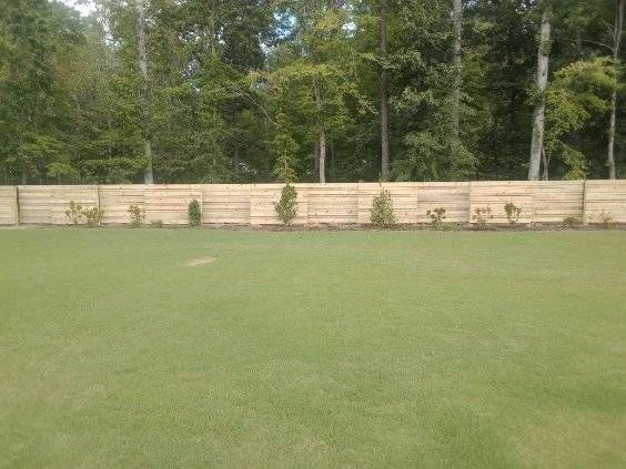 A wide, lush green lawn stretches toward a horizontal wooden fence backed by a line of mature trees.