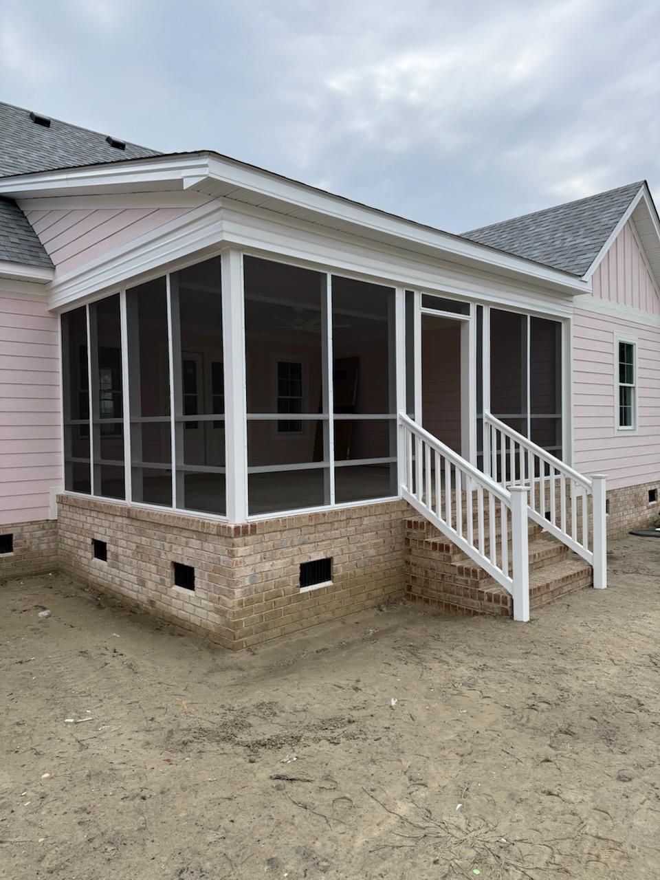 A pink house with a screened-in porch, brick foundation, white trim, and white stairs on a dirt lot.