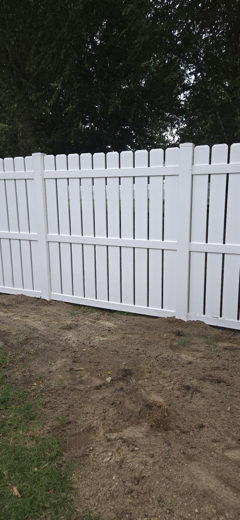 A white vinyl privacy fence with vertical pickets stands in a dirt yard with trees in the background.