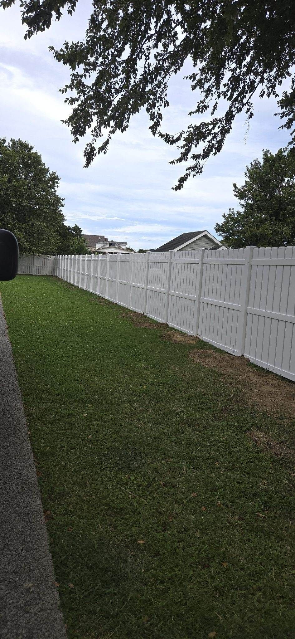A white vinyl fence runs along the side of a grassy residential backyard under a cloudy sky.