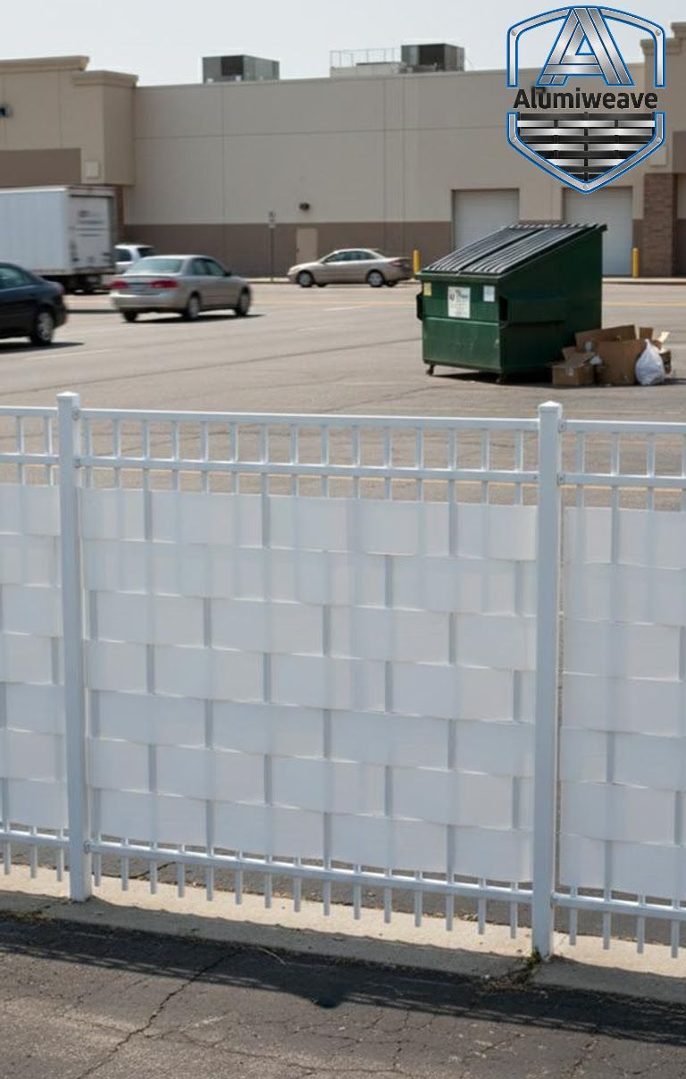 A white aluminum fence with privacy slats installed in a commercial parking lot near a dumpster.