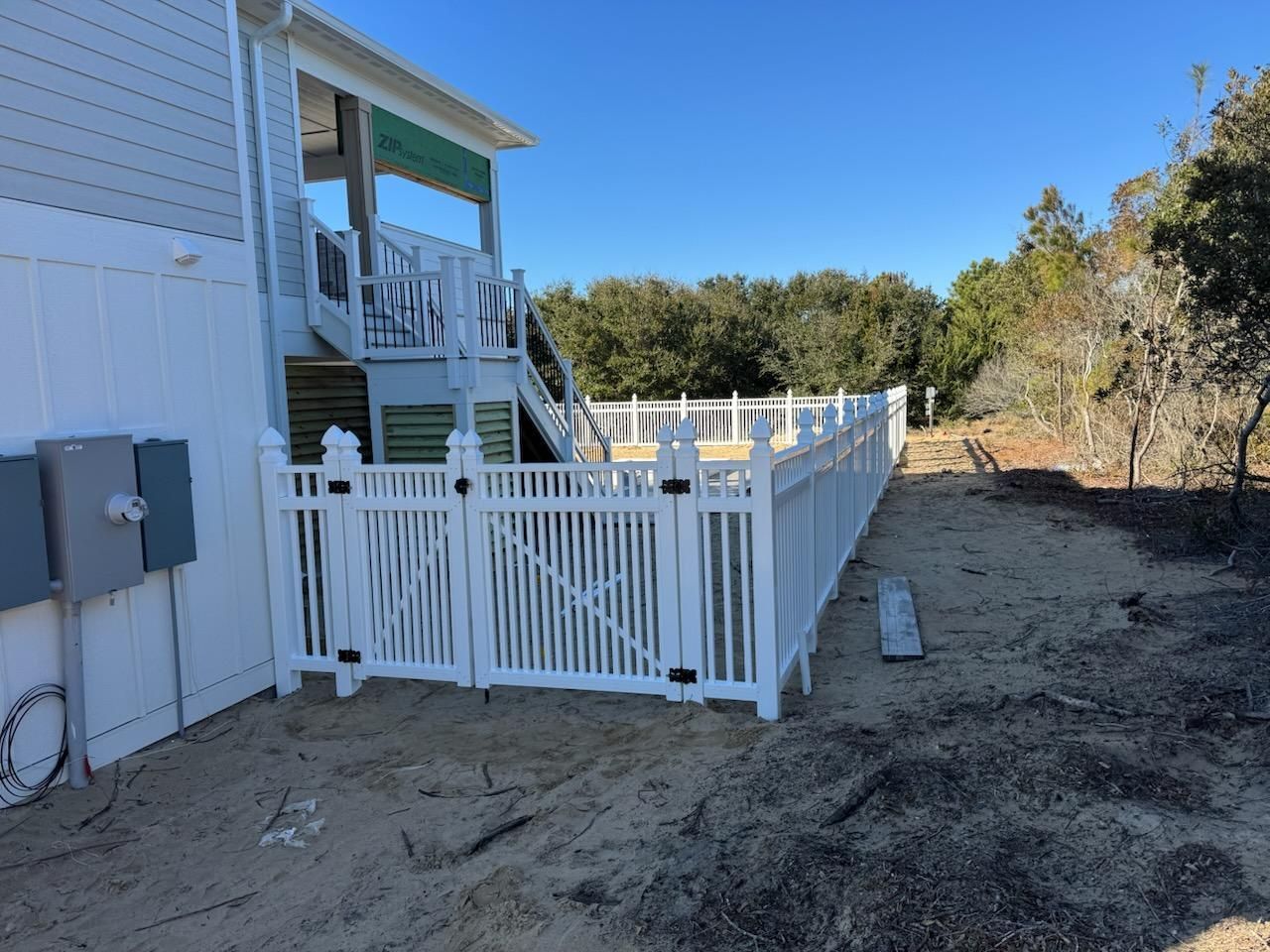 A white vinyl picket fence with a double gate sits beside a house on a sandy lot with woods in the background.