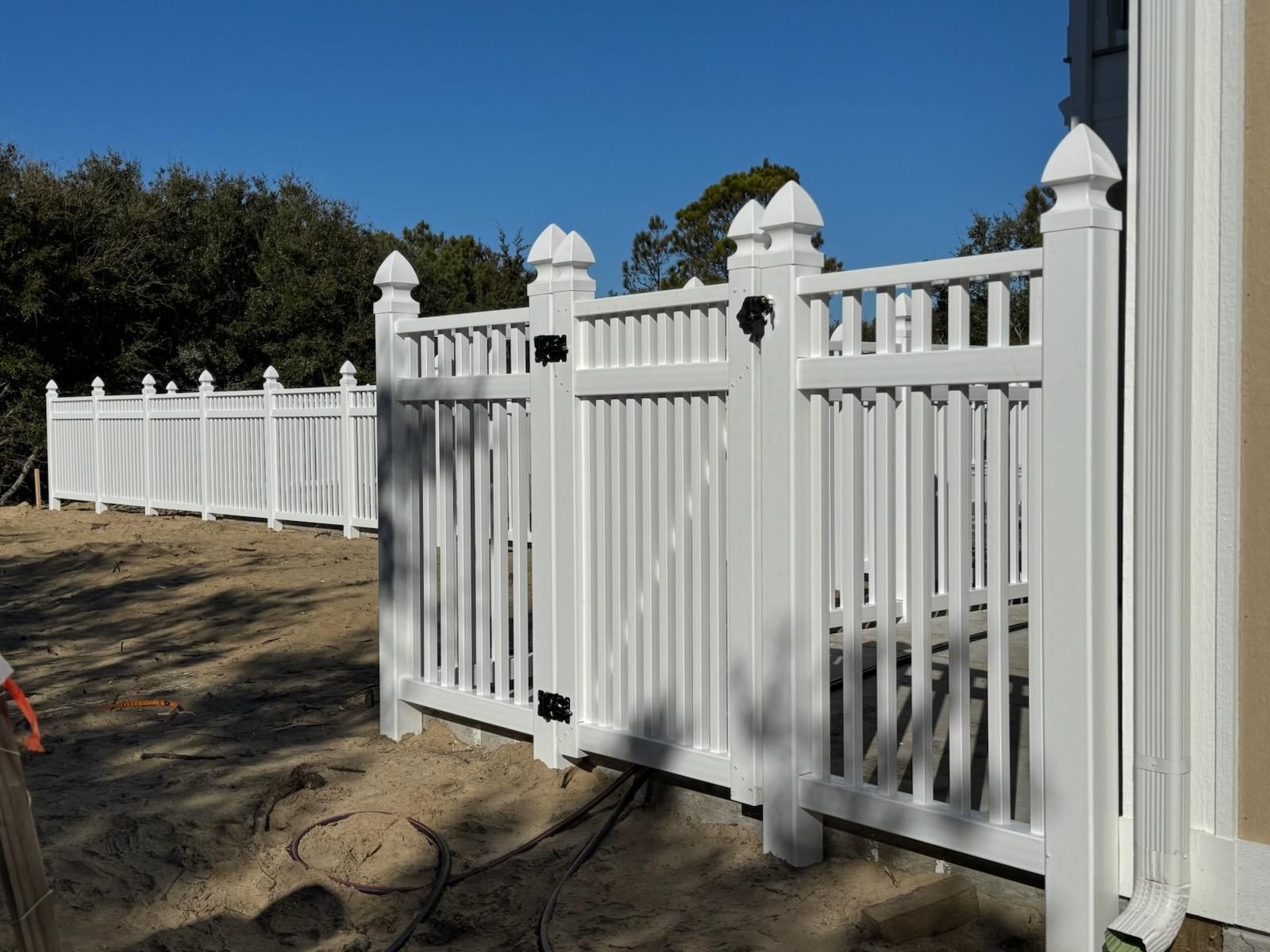 A white vinyl fence with a matching gate, installed on a sandy plot of land next to a tan house exterior.