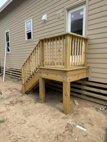 A newly constructed wooden deck and staircase attached to the side of a light-brown house on a sandy, unfinished lot.