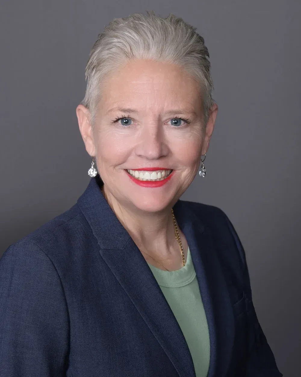 Woman with short gray hair, smiling, wearing a blue blazer, light green shirt, and pearl earrings against a gray backdrop.