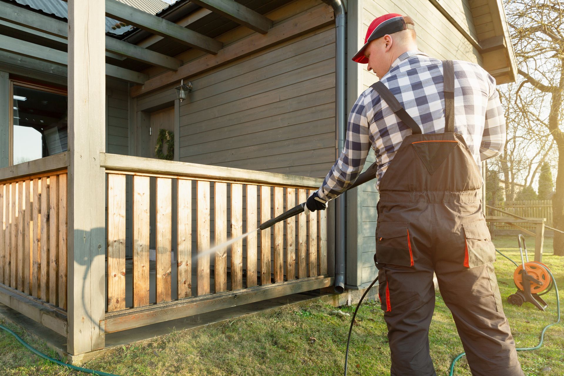 Man power washing a wooden railing on a house, wearing overalls and a red cap.