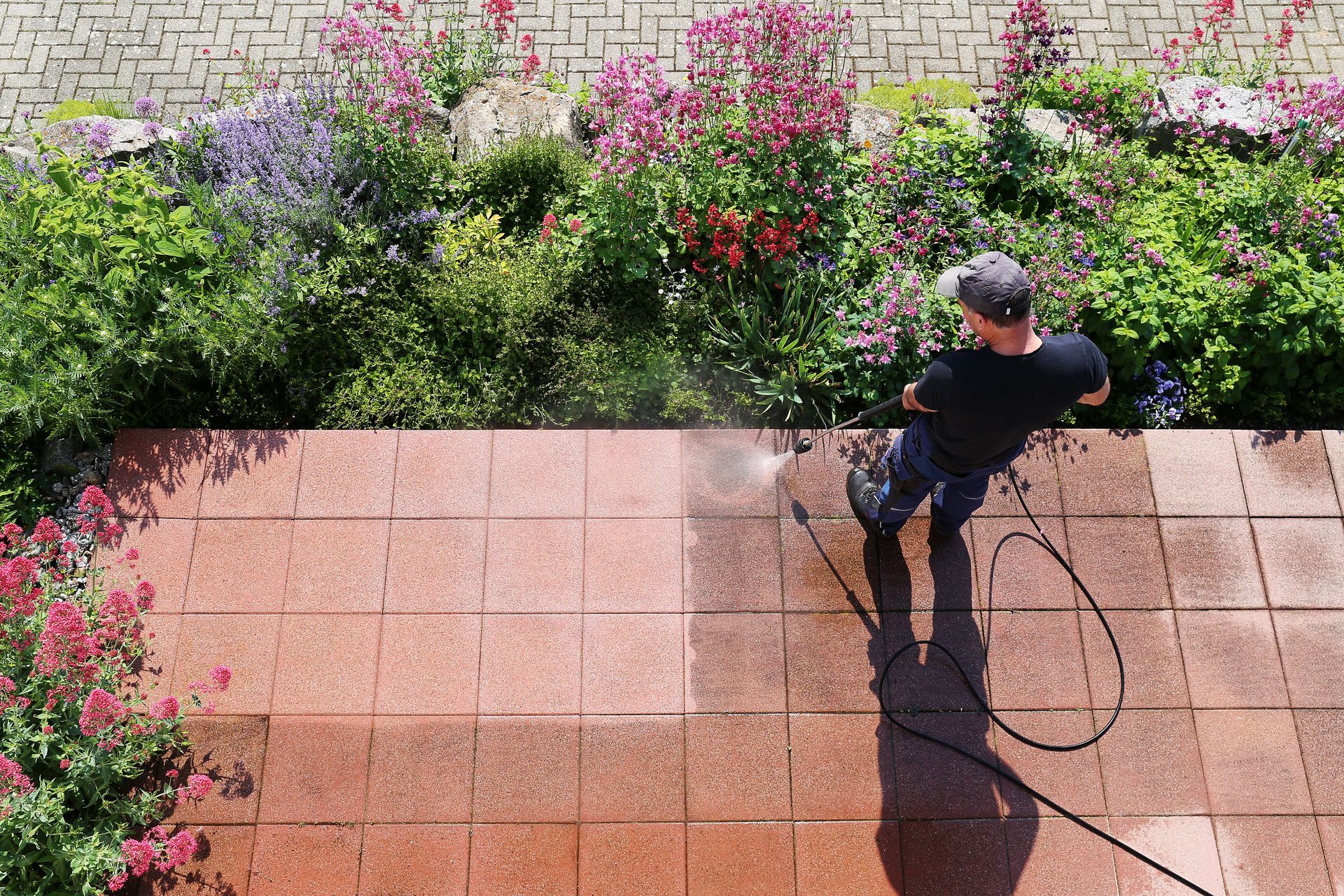 Person pressure washing red patio next to a colorful flower garden.