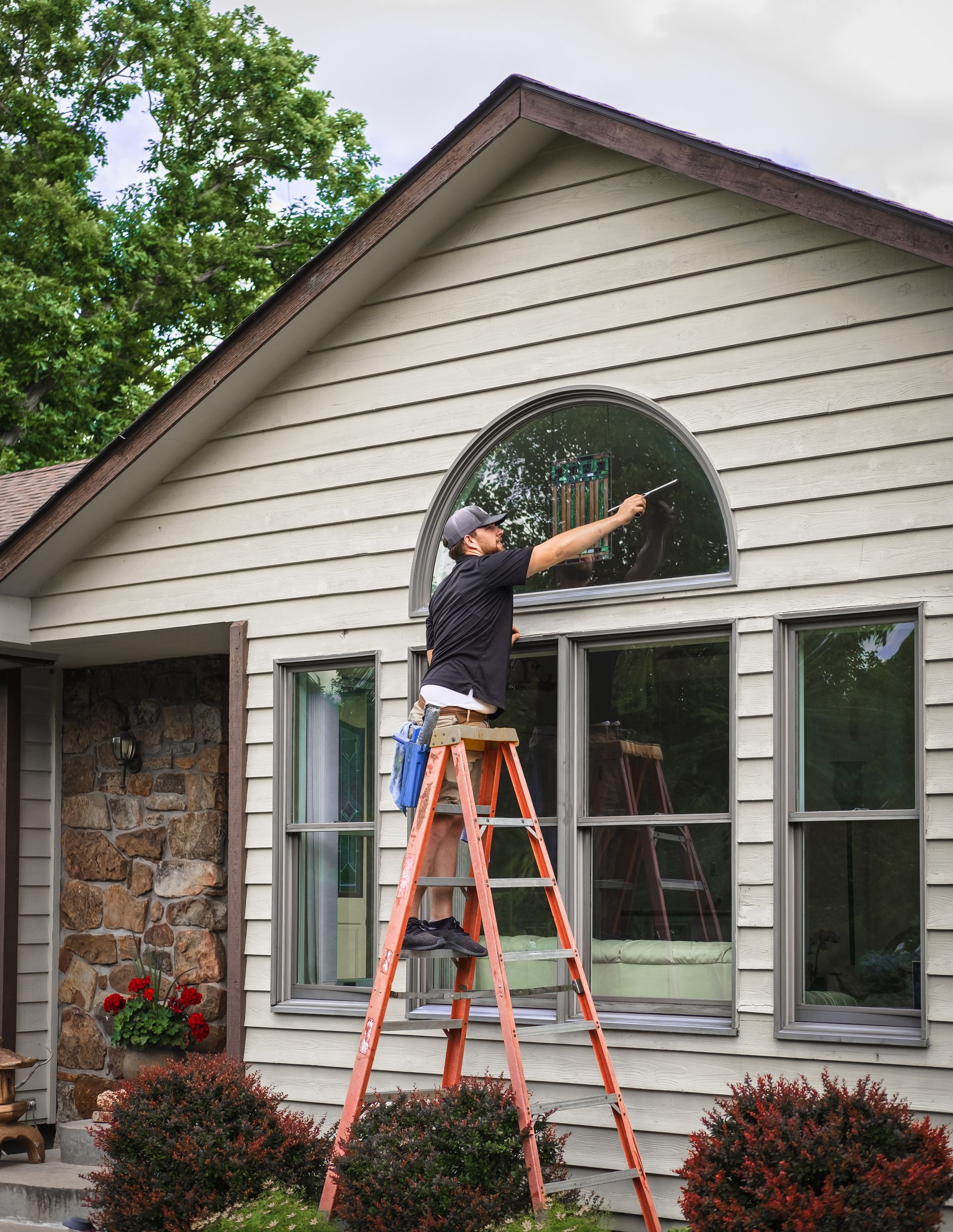 Window cleaner on ladder washing arched window of a beige house.