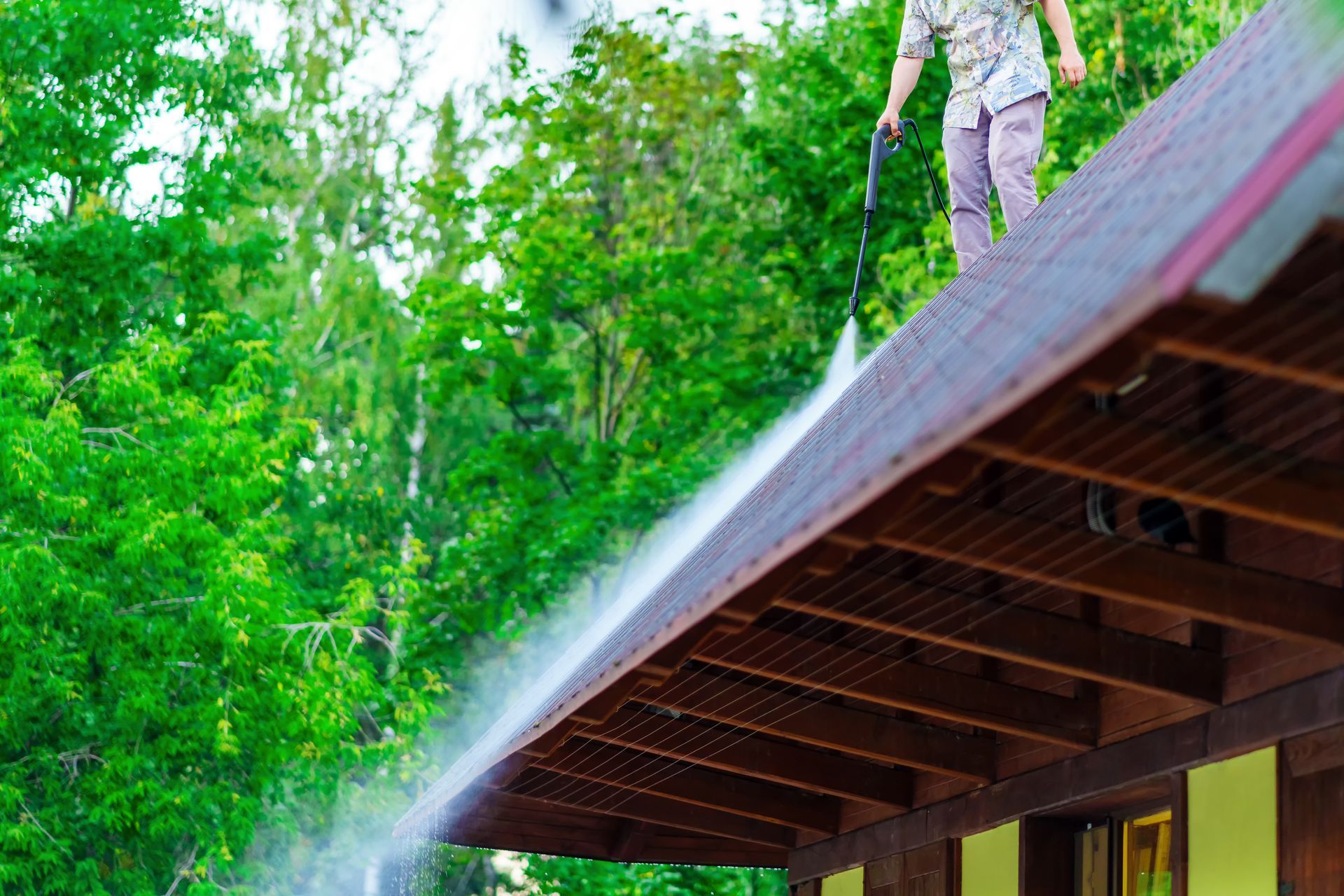 Person pressure washing a brown roof with a water spray. Green trees in the background.