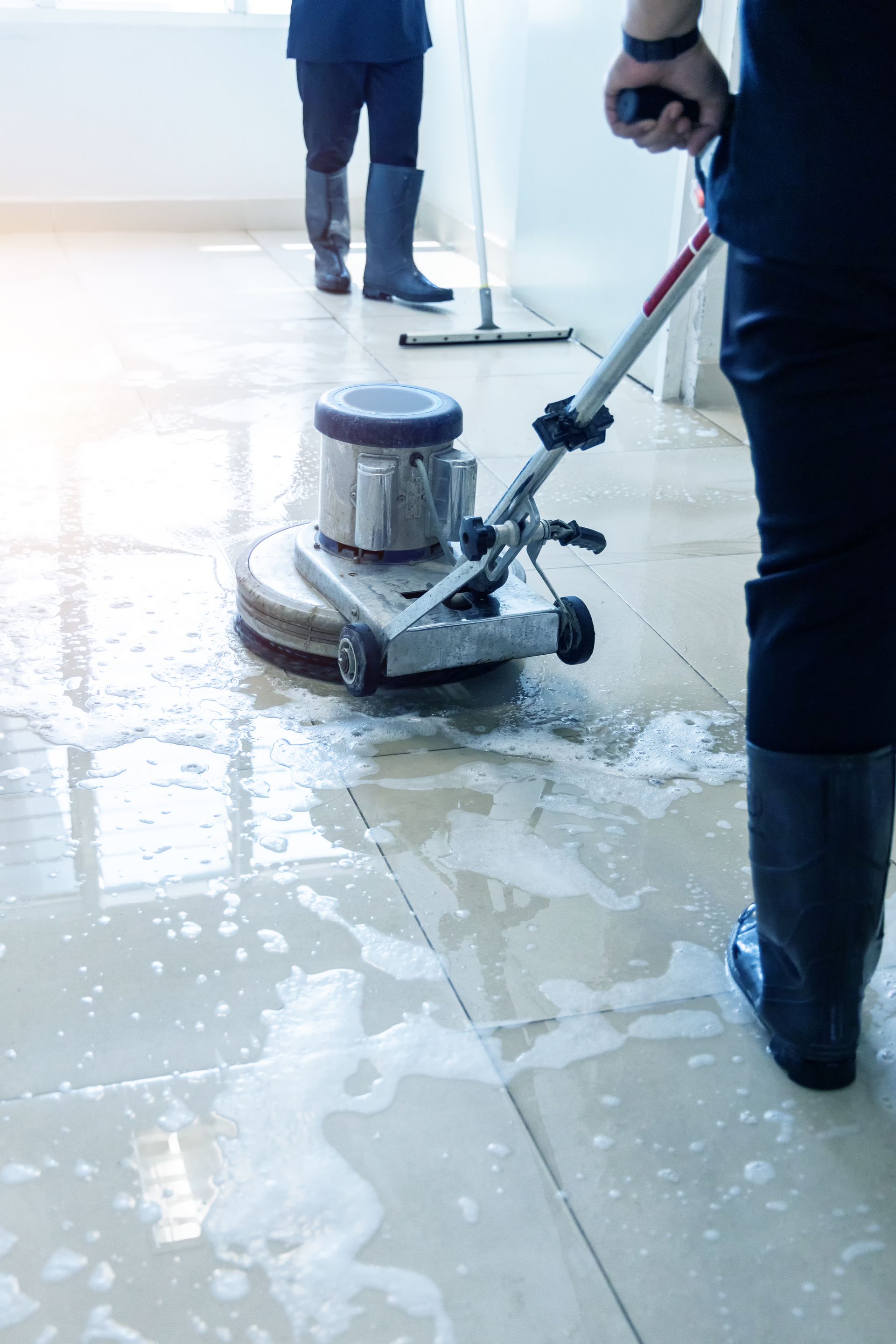 Person using floor cleaning machine. Wet floor, suds, and another person in background with a mop.
