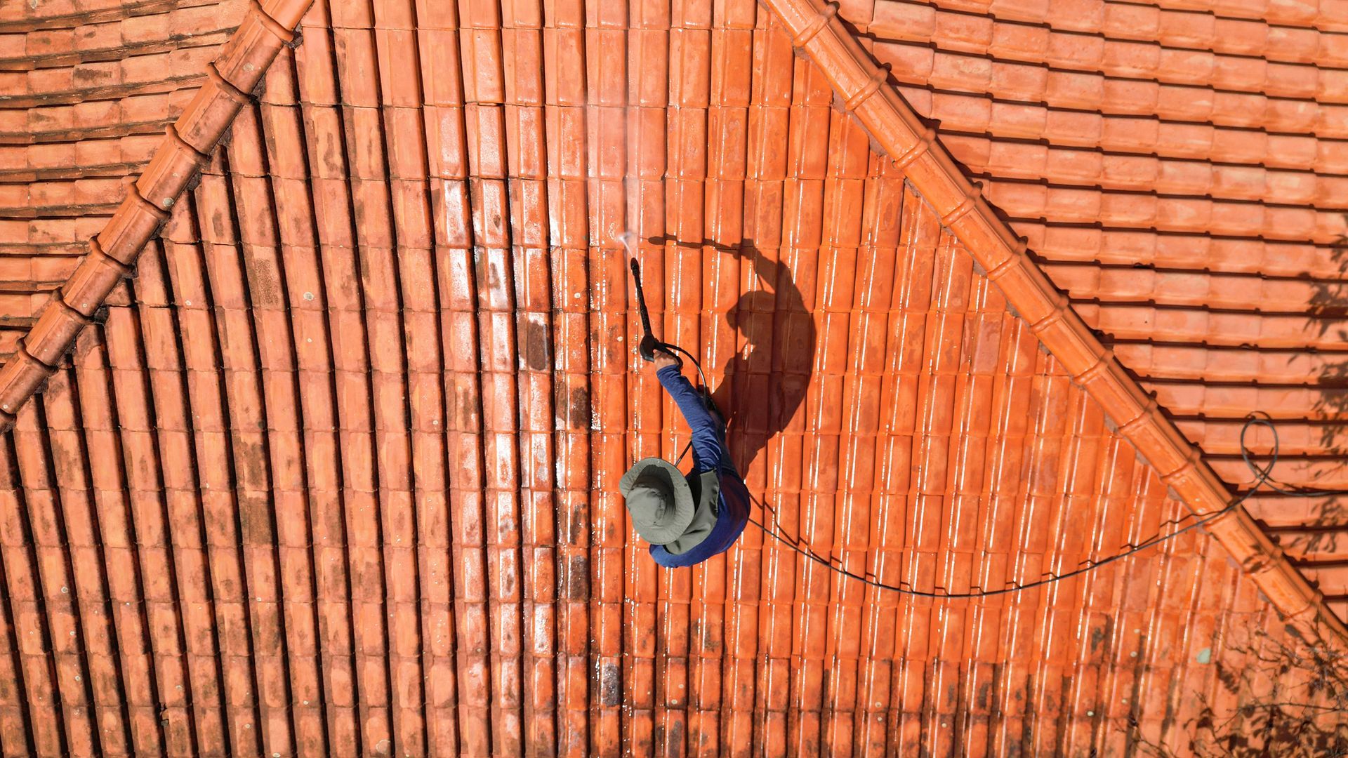 Person on a roof spraying water, cleaning terracotta tiles. Overhead view with the sun casting a long shadow.