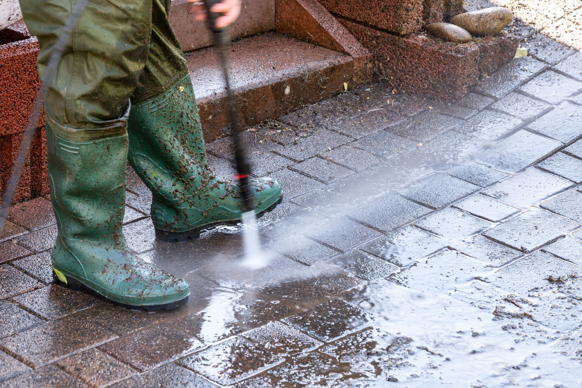 Person in green rubber boots power washing brick patio.