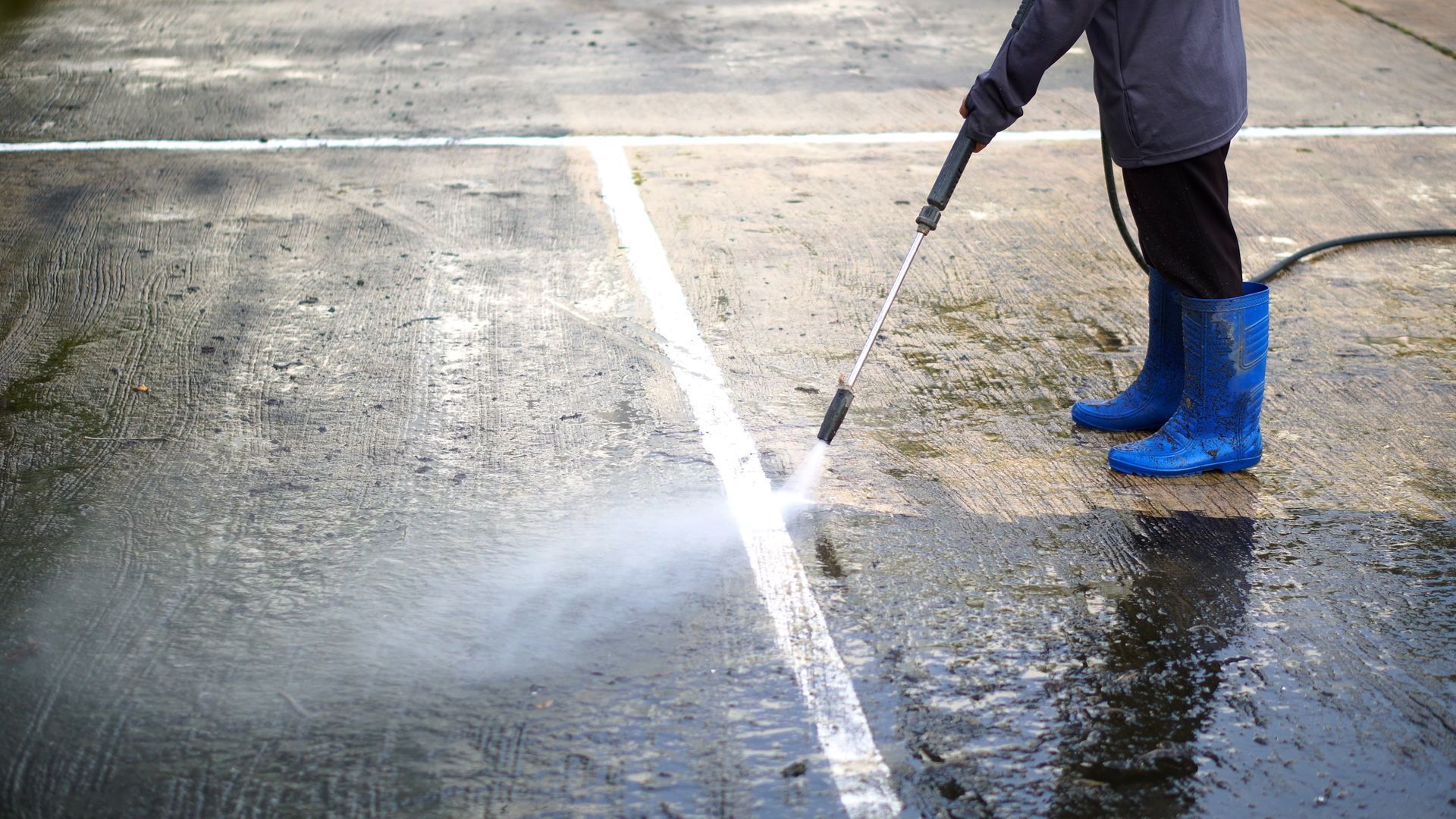 Person power washing a concrete surface, removing dirt and revealing a white line.