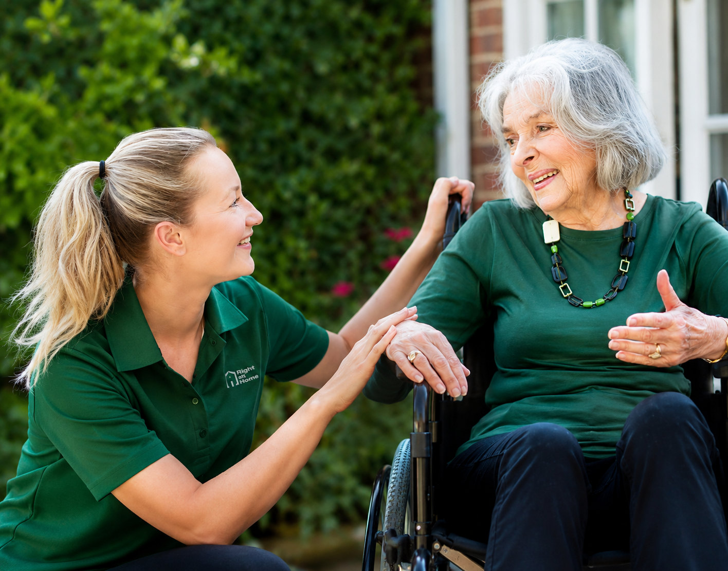 Caregiver in green scrubs smiles while talking with an older woman in a wheelchair outdoors.