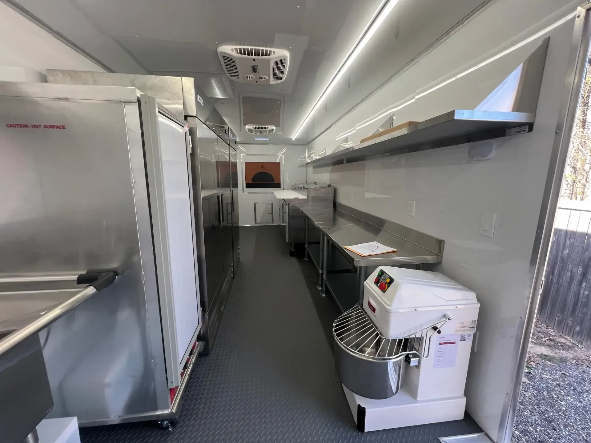 Interior of a commercial kitchen trailer featuring stainless steel surfaces, a mixer, and refrigerated units along the walls