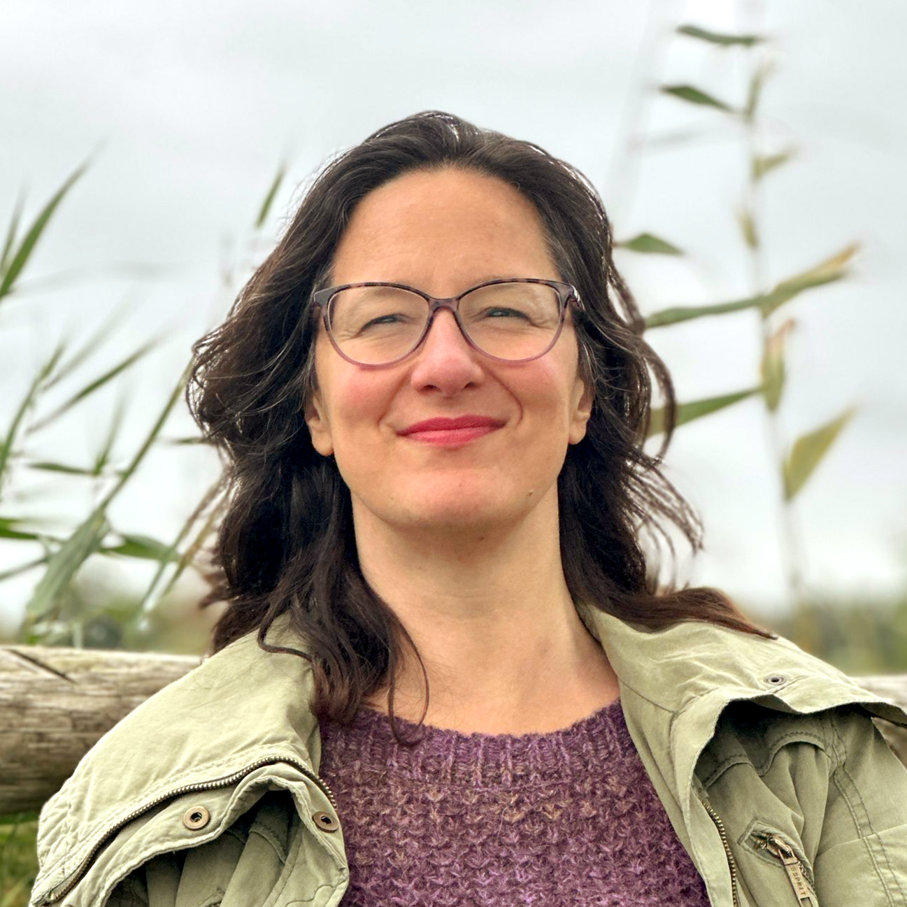 Woman with glasses smiles outdoors, wearing a purple sweater and green jacket, with natural backdrop.