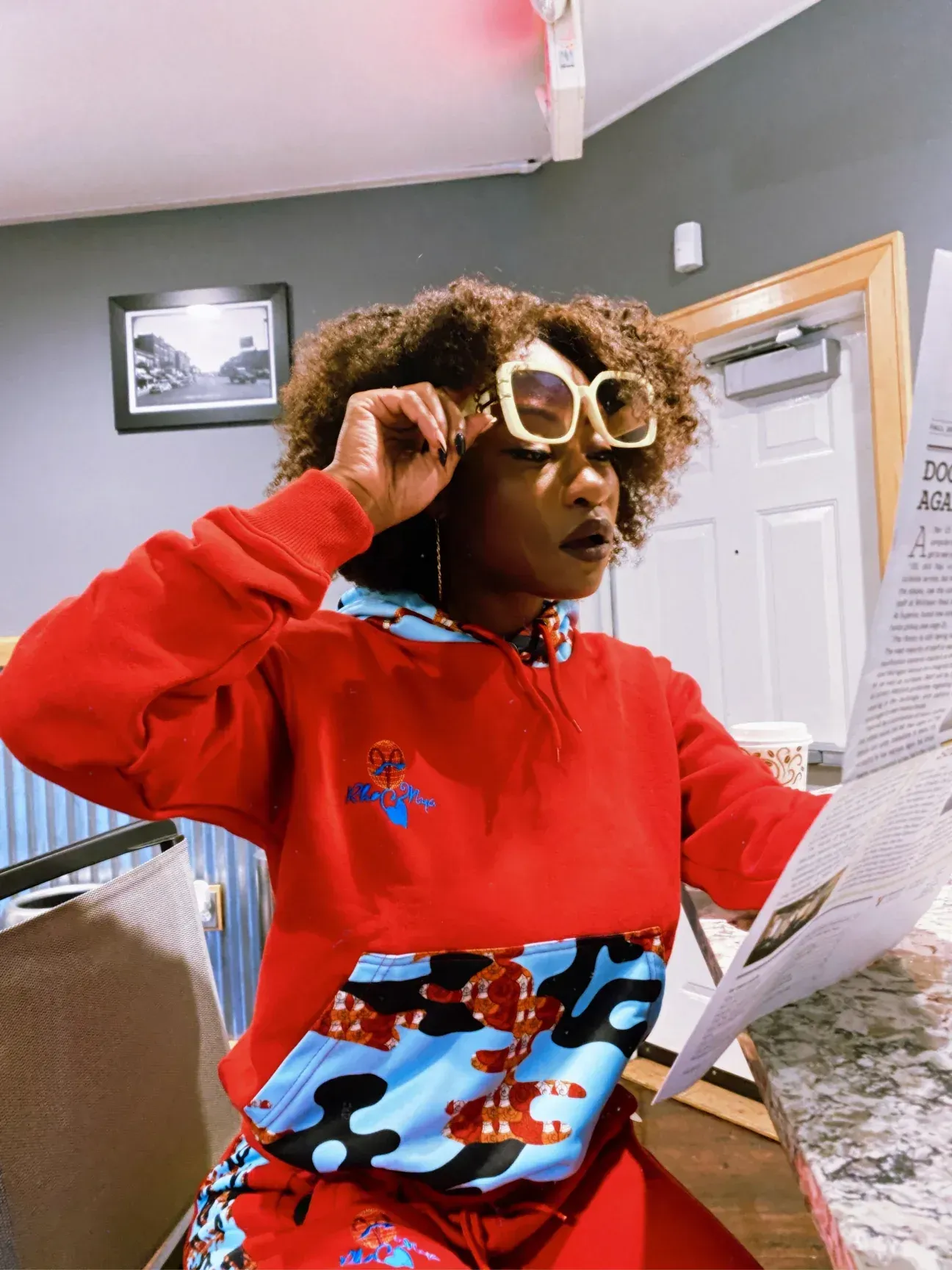 A woman wearing sunglasses and a red hoodie is reading a newspaper.