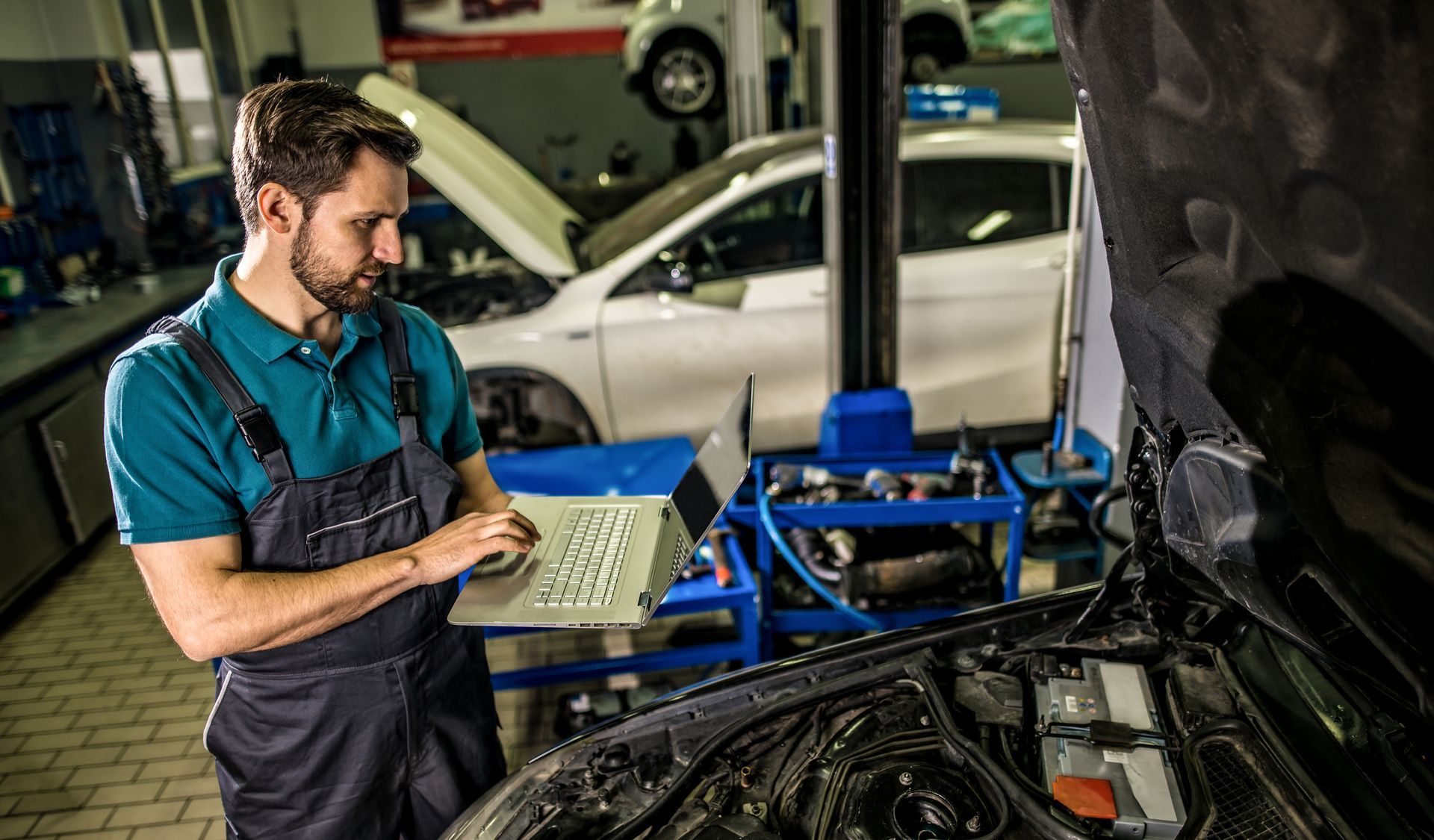 Mechanic in blue overalls using a laptop to diagnose a car engine in a garage.