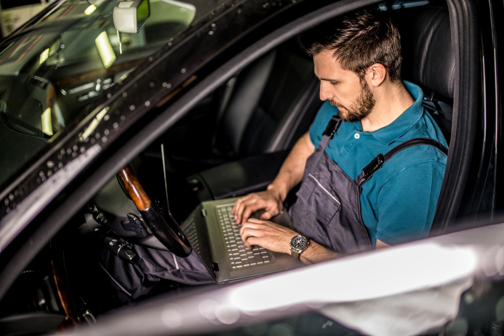 Mechanic in overalls uses a laptop inside a car for diagnostics.