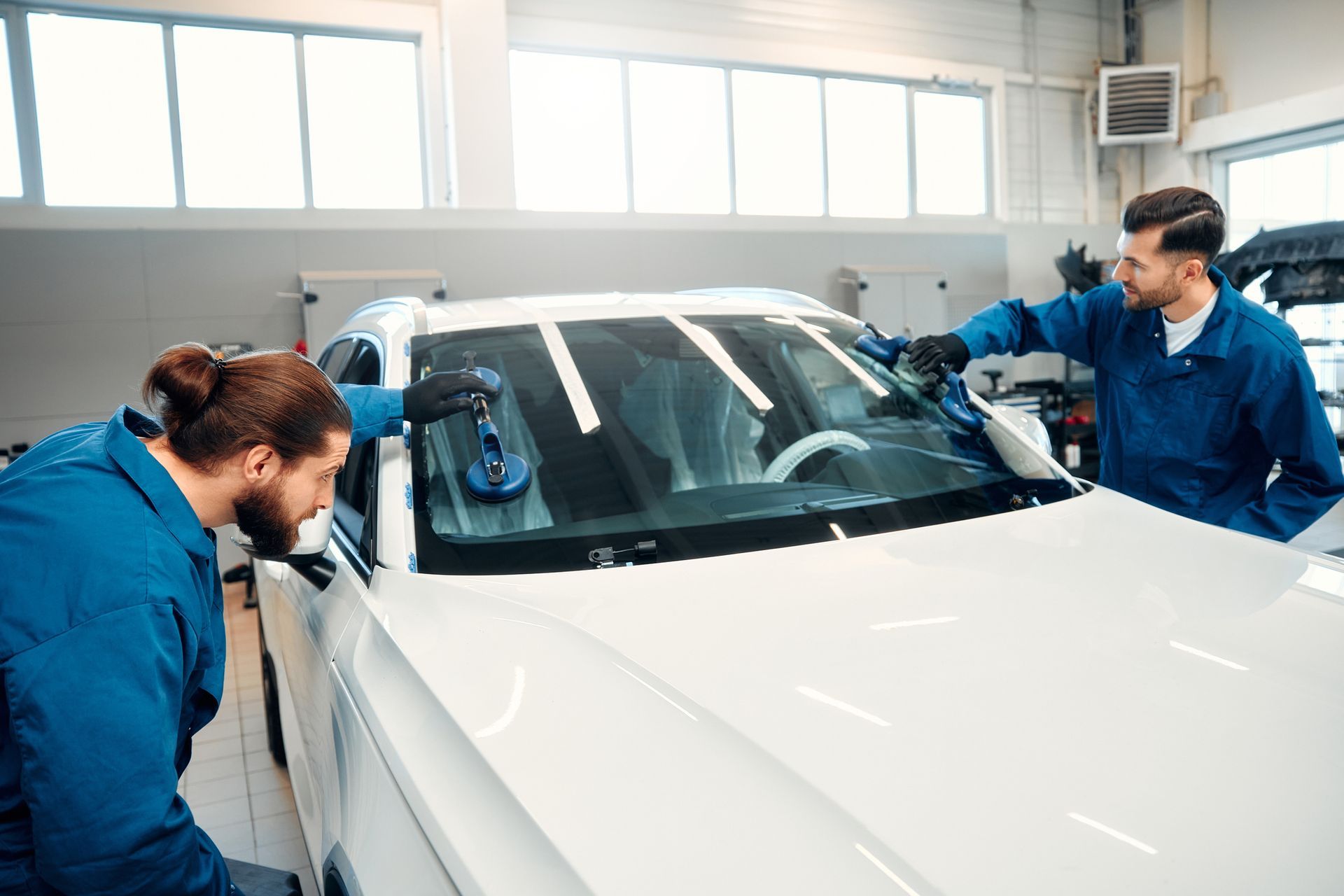 Two men in blue coveralls install a windshield in a white car inside a garage.